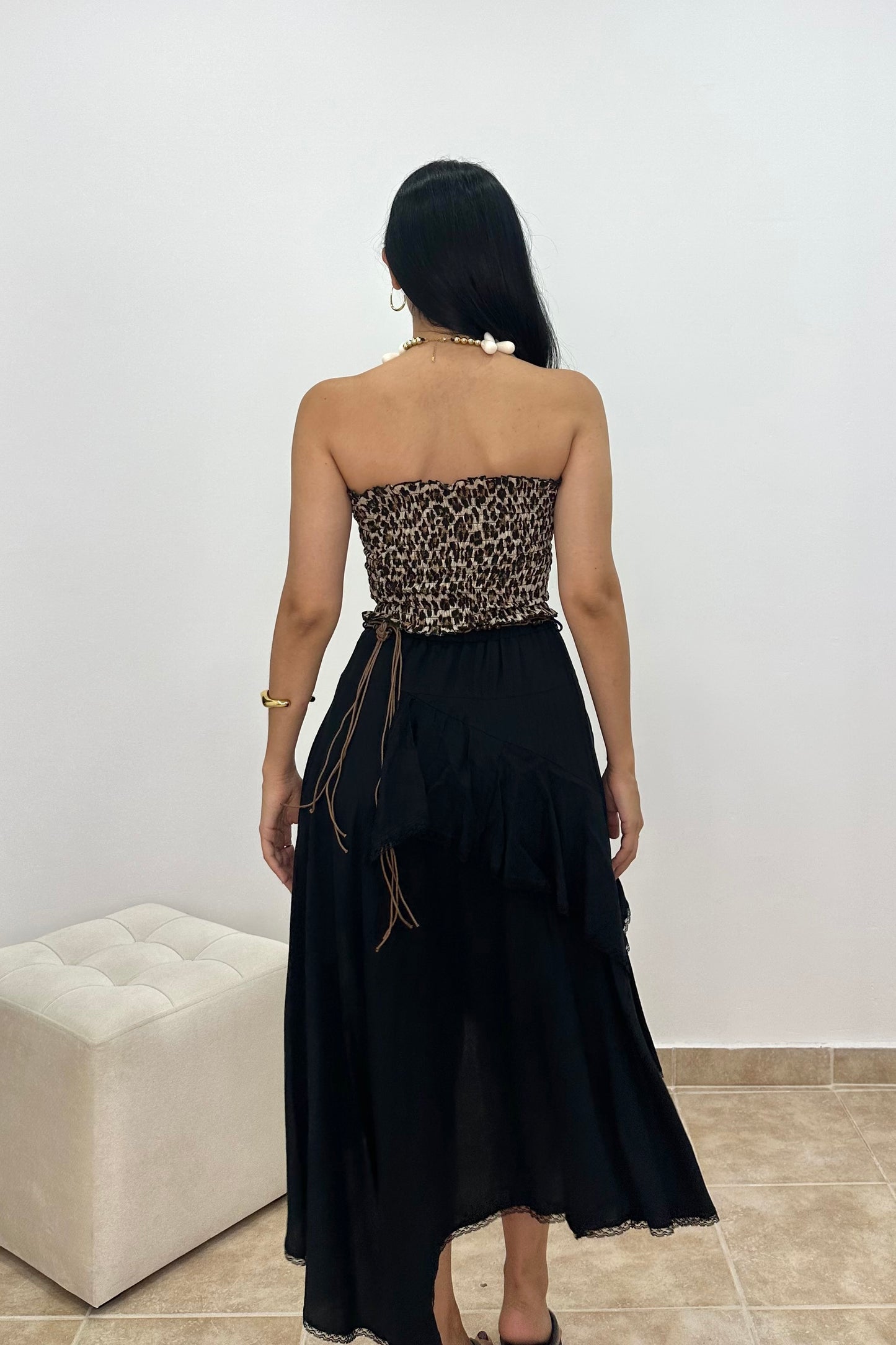A woman viewed from behind wearing a strapless black dress with a leopard-print bandeau-style bodice and a long black skirt, standing indoors against a plain light wall.