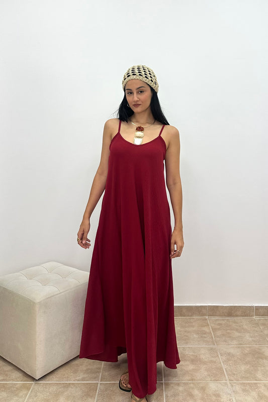 A woman modeling a long, sleeveless burgundy maxi dress with thin spaghetti straps and a scoop neckline, styled with a woven headband and a gold pendant necklace, standing in front of a white wall.