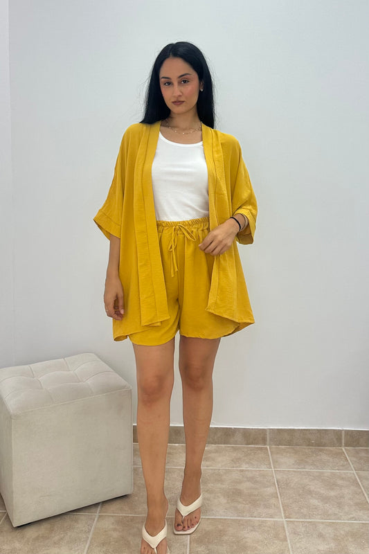 A woman modeling a matching yellow two-piece lounge set consisting of a short-sleeve open-front cardigan and drawstring shorts, worn over a white tank top and paired with white sandals.