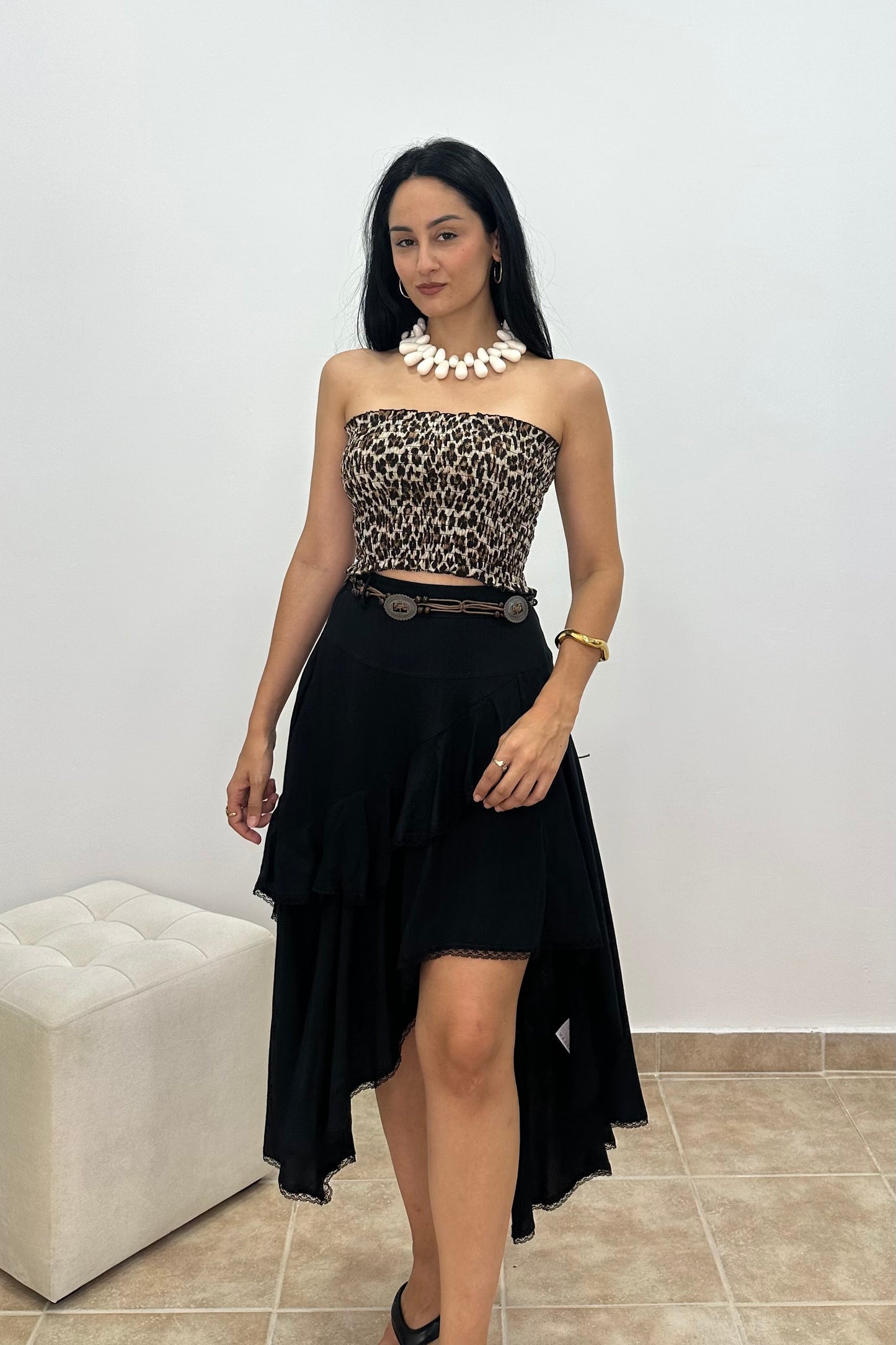 A woman modeling a strapless black and brown leopard-print crop top paired with a high-low black skirt and a black belt with silver hardware, accessorized with a chunky white collar necklace.