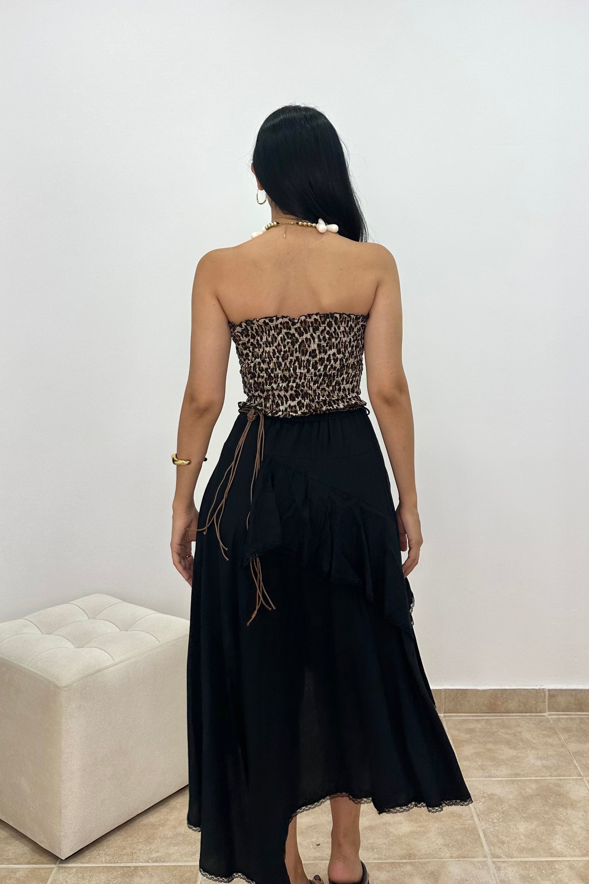A woman viewed from behind wearing a strapless black dress with a leopard-print bandeau-style bodice and a long black skirt, standing indoors against a plain light wall.
