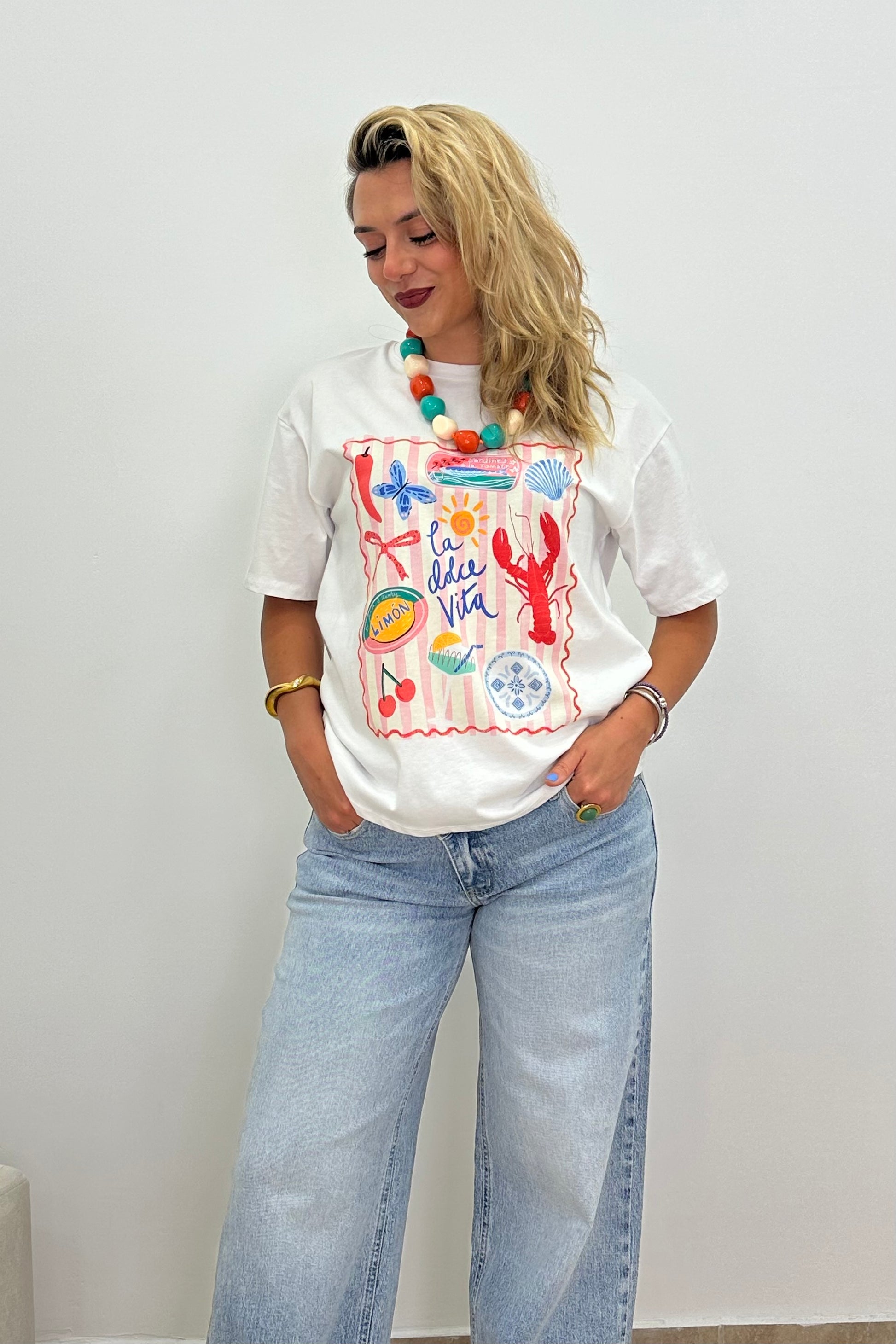 A woman wearing a white graphic t-shirt with a colorful ocean-themed print, paired with light-wash wide-leg jeans and a chunky beaded necklace, standing against a plain white wall.