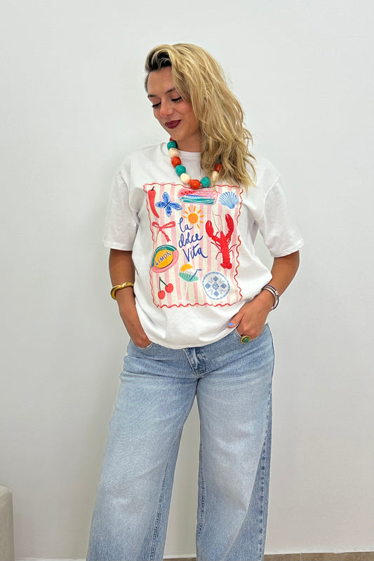 A woman wearing a white graphic t-shirt with a colorful ocean-themed print, paired with light-wash wide-leg jeans and a chunky beaded necklace, standing against a plain white wall.