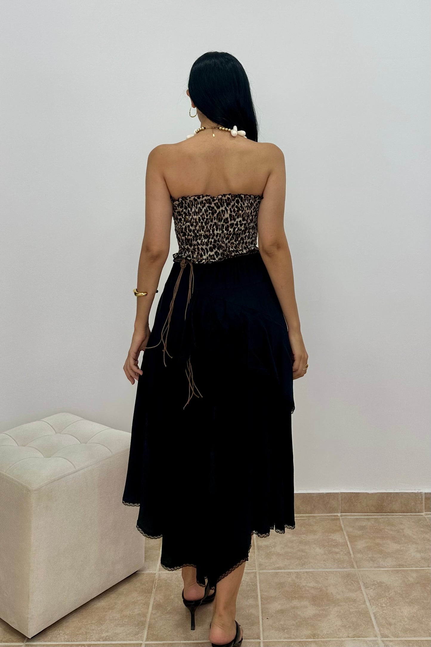 A woman is shown from behind wearing a strapless black dress with a leopard-print patterned bodice and a long black skirt, standing in an indoor setting with a tiled floor and a white tufted bench.