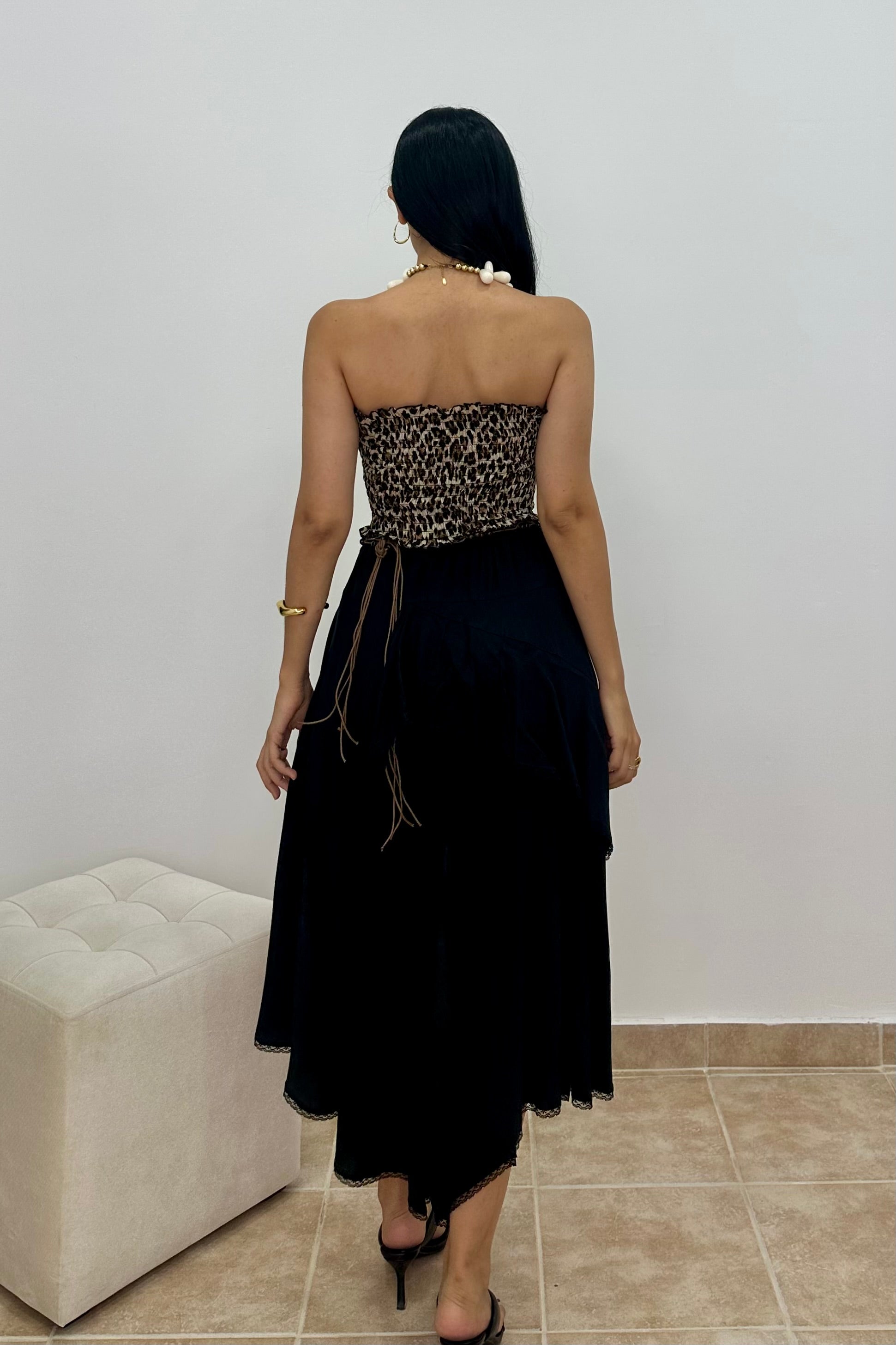 A woman is shown from behind wearing a strapless black dress with a leopard-print patterned bodice and a long black skirt, standing in an indoor setting with a tiled floor and a white tufted bench.