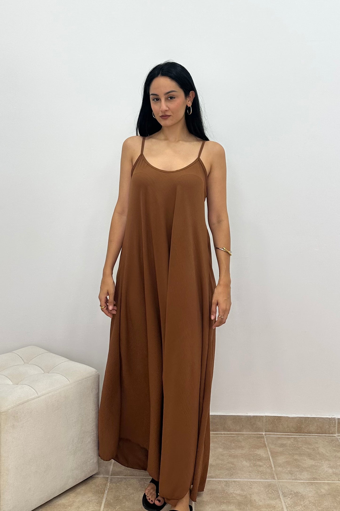 A woman modeling a brown maxi slip dress with thin spaghetti straps, standing in front of a plain white wall.