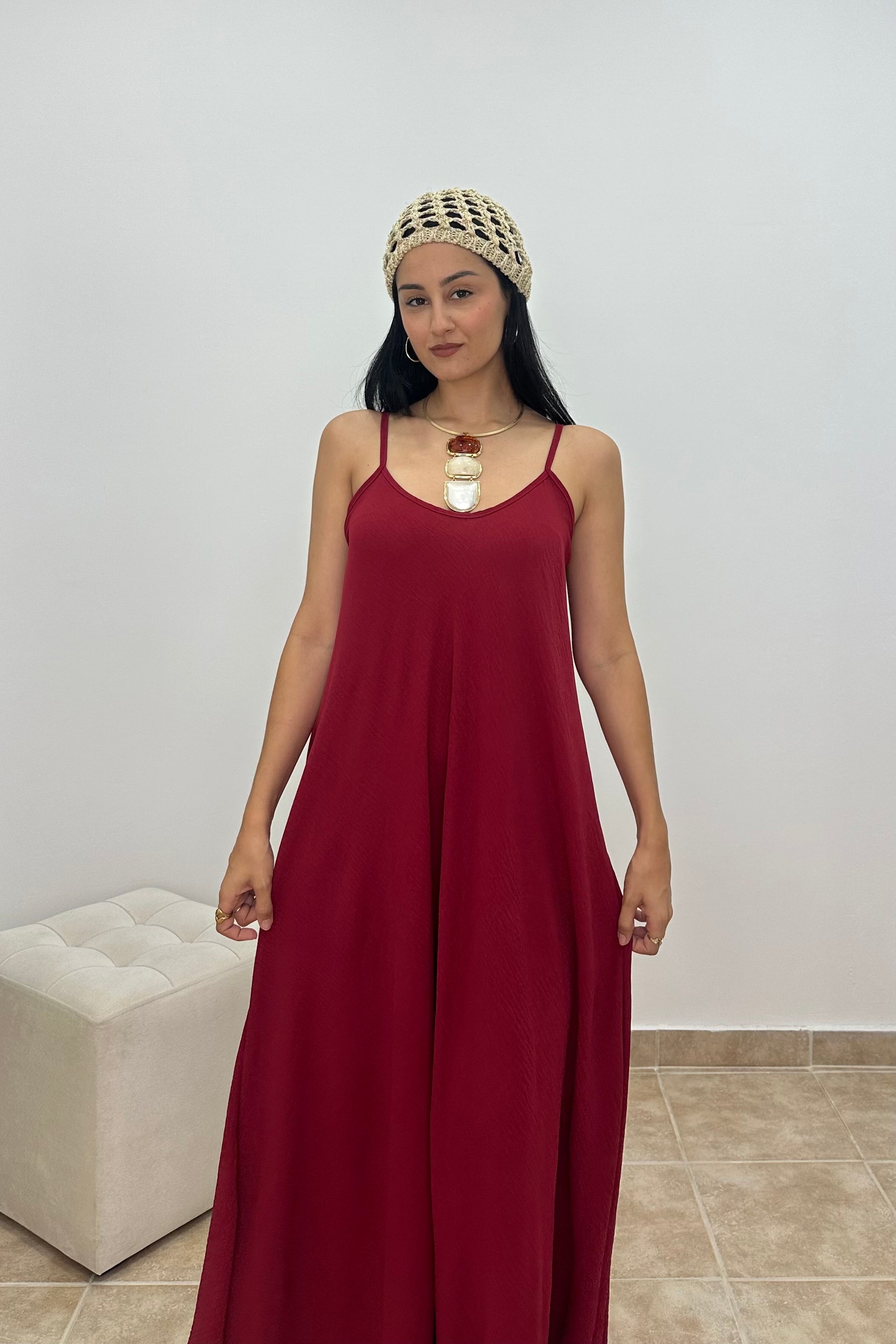 A woman wearing a long, sleeveless burgundy maxi dress with thin straps, a chunky knit beige headband, and a large pendant necklace, standing in a minimalist room with a white wall and a tufted ottoman.