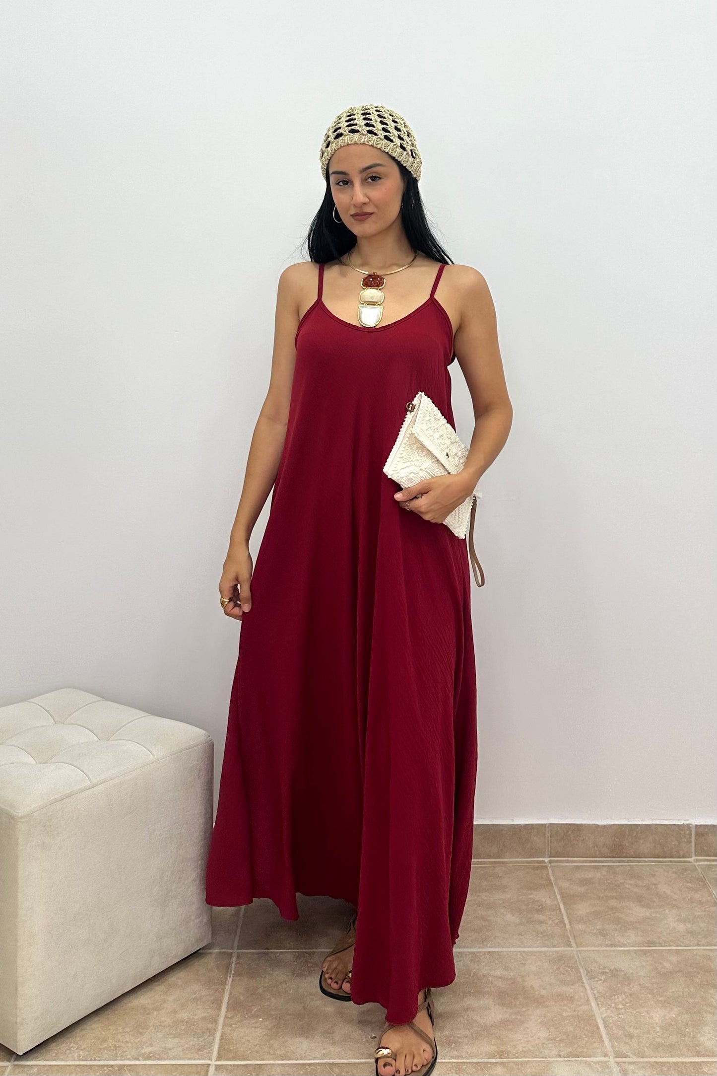 A woman modeling a sleeveless, floor-length burgundy maxi dress with thin straps, standing indoors against a plain light gray wall and holding a small white clutch.