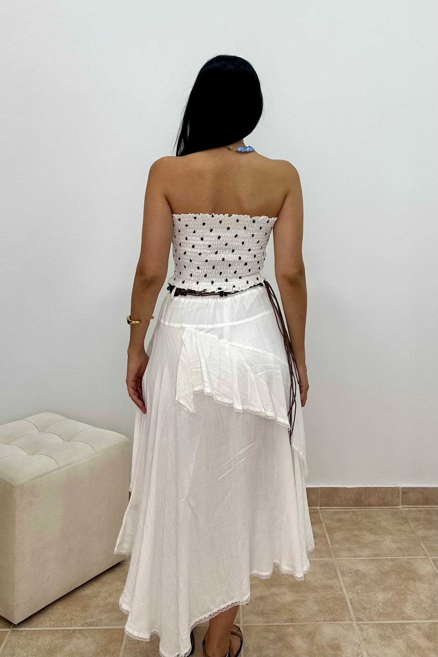 A woman viewed from behind wearing a white strapless dress with a black polka-dot bodice, a thin black waist tie, and a layered ruffled white skirt, standing in an indoor room with a light wall and tiled floor.