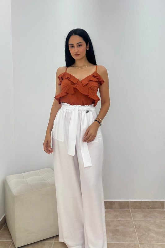 A woman modeling a two-piece outfit consisting of a rust-orange ruffled spaghetti-strap crop top and high-waisted white wide-leg pants with a tie waist, standing in a minimalist room with a white wall and a beige ottoman.