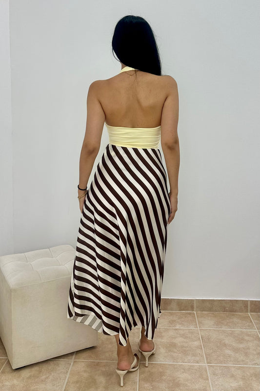 A woman viewed from behind wearing a strapless halter maxi dress with a cream and dark brown diagonal stripe pattern and a solid cream waistband. She is standing in a room with a light-colored wall, tiled floor, and a tufted ottoman.