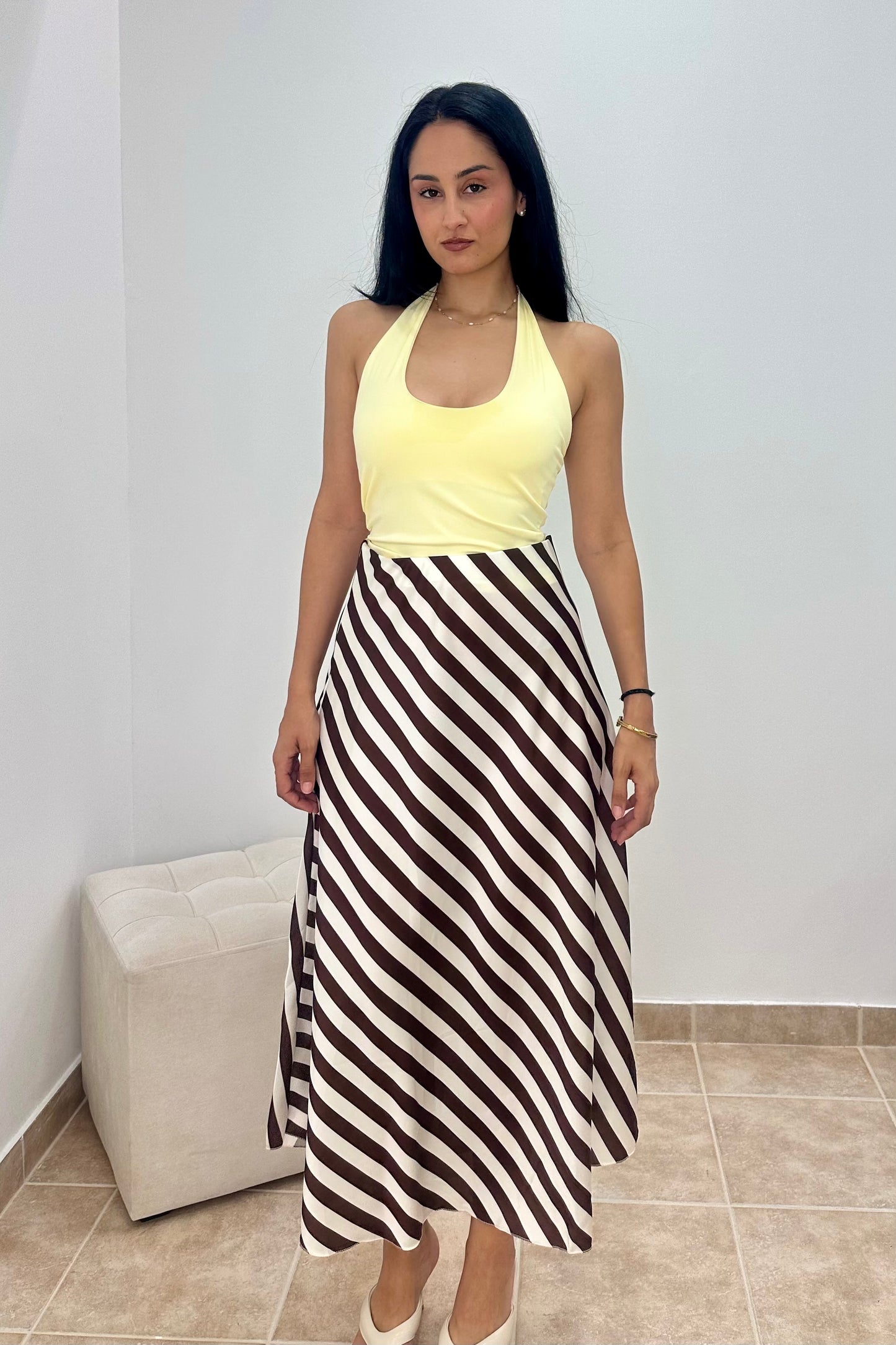 A woman modeling a sleeveless yellow halter top paired with a brown and white diagonally striped midi skirt, standing in a minimalist room with a white wall and tiled floor.