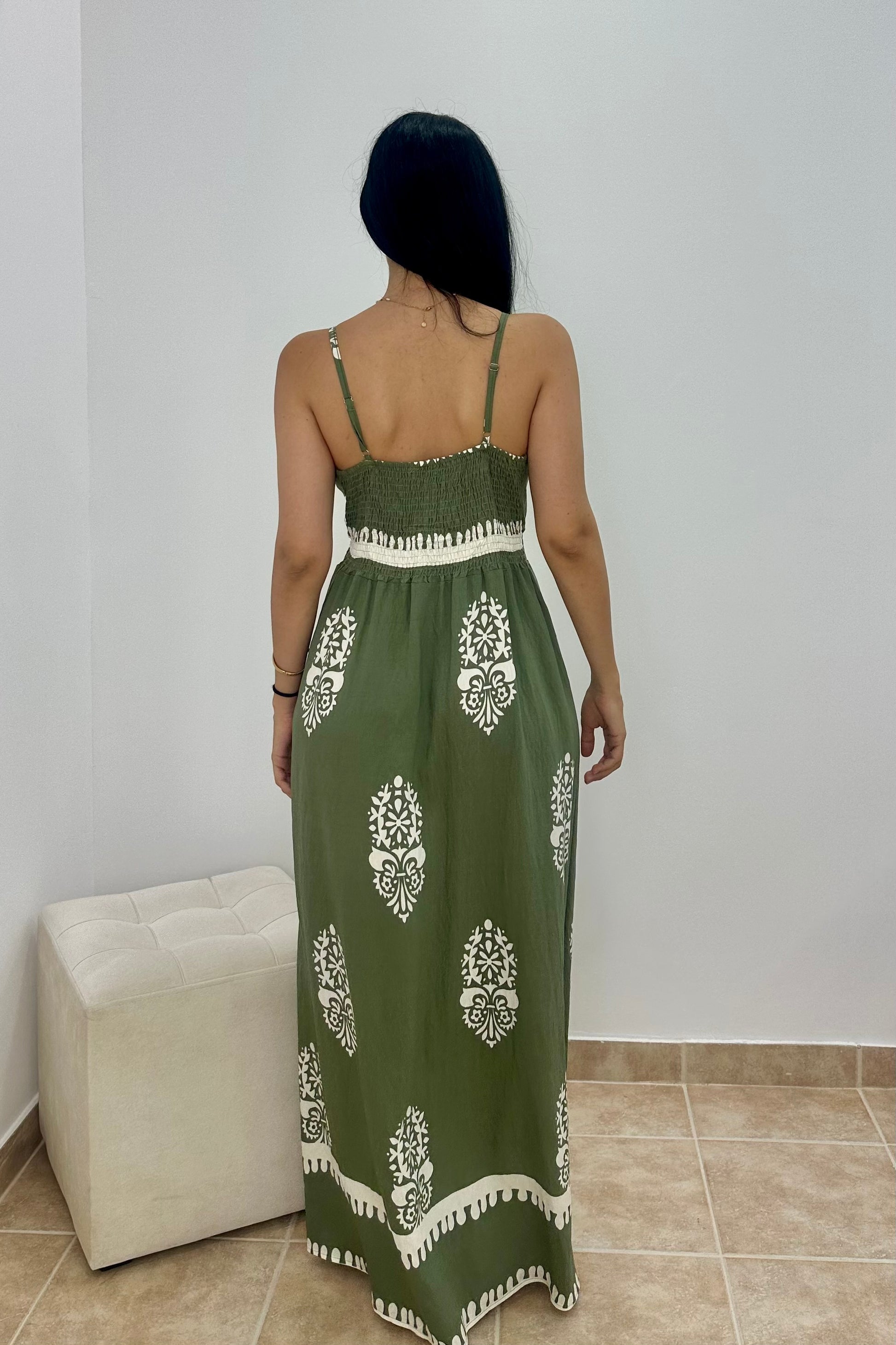 A woman viewed from behind wearing a sleeveless olive green maxi dress with white printed motifs and a white tassel-trimmed hem, standing indoors against a plain light wall.