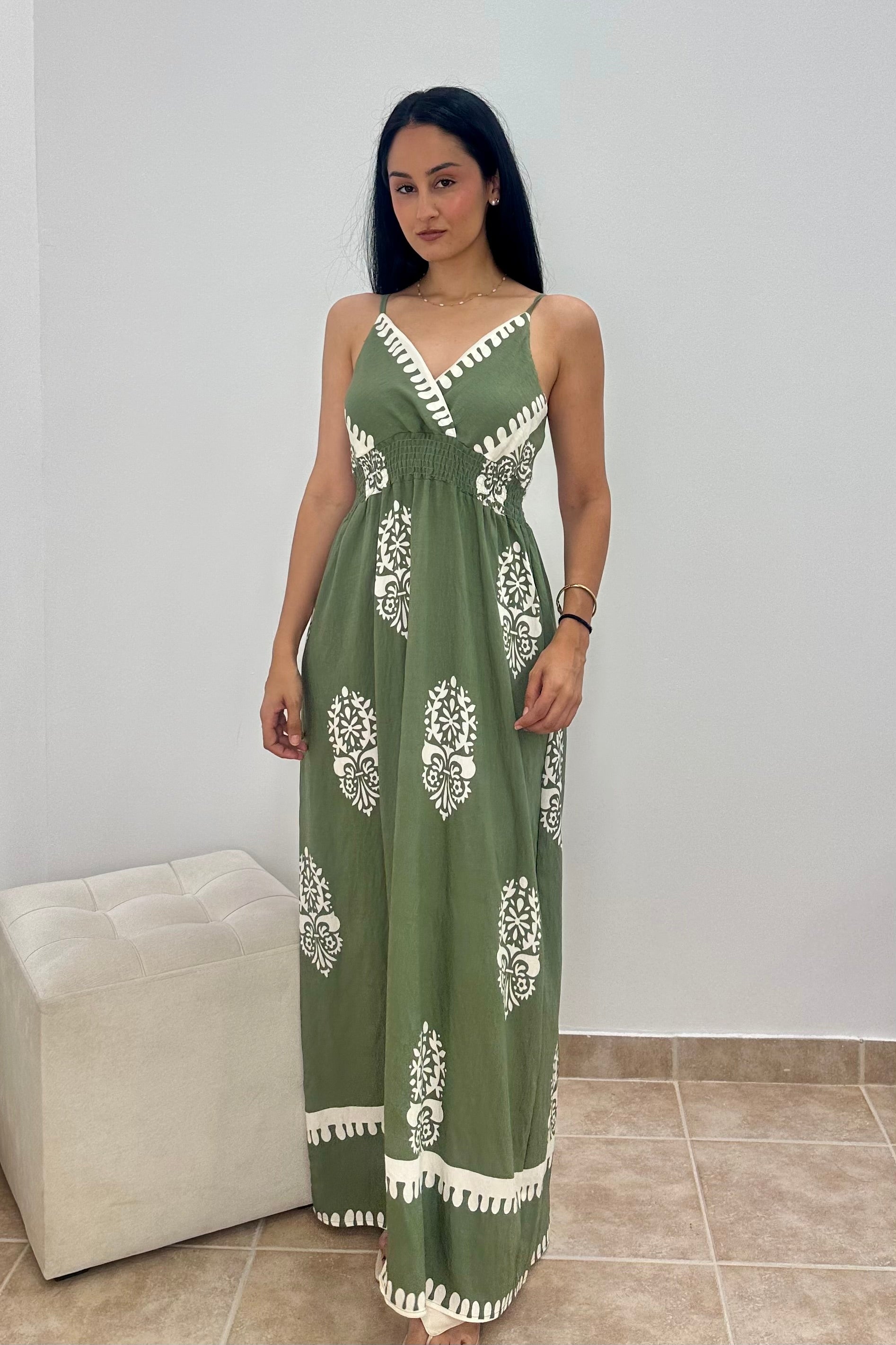 A woman modeling a green maxi dress with white printed motifs and white trim details, standing in an indoor setting with a light-colored wall and a white ottoman.