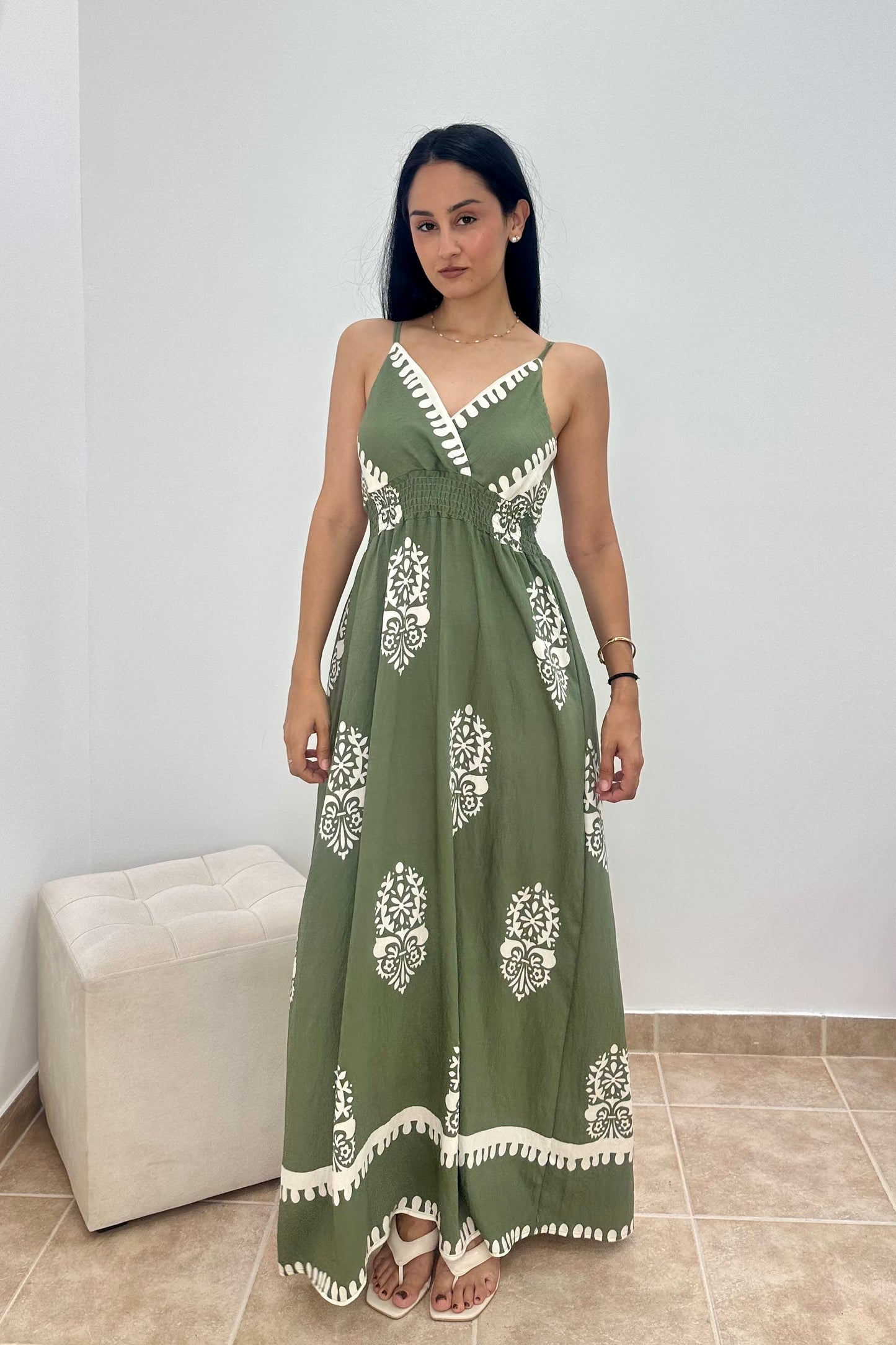 A woman modeling a sleeveless olive green maxi dress with white block prints and a white tassel-trimmed neckline, standing in a minimalist room with a white wall and beige floor tiles.