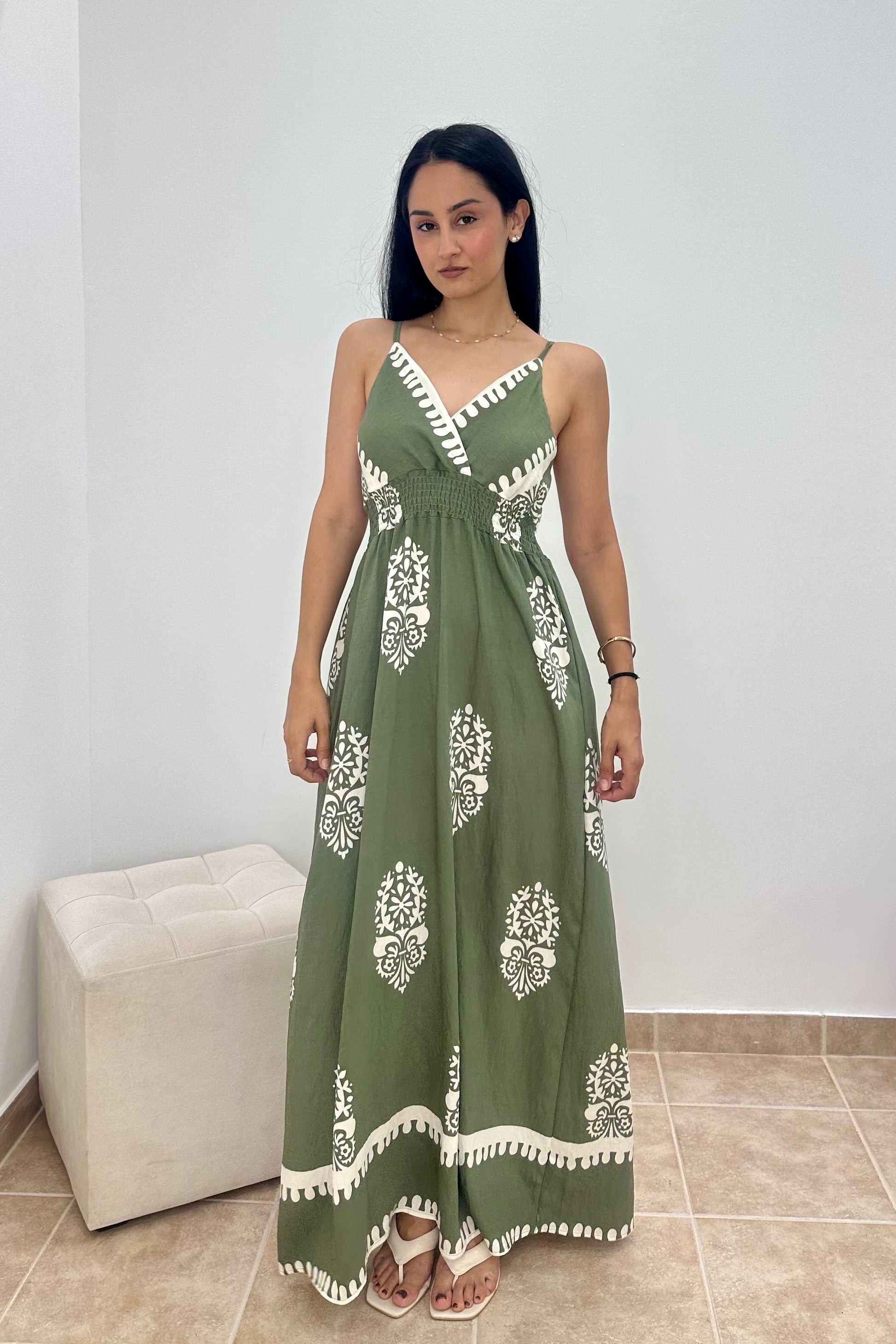 A woman modeling a sleeveless olive green maxi dress with white block prints and a white tassel-trimmed neckline, standing in a minimalist room with a white wall and beige floor tiles.