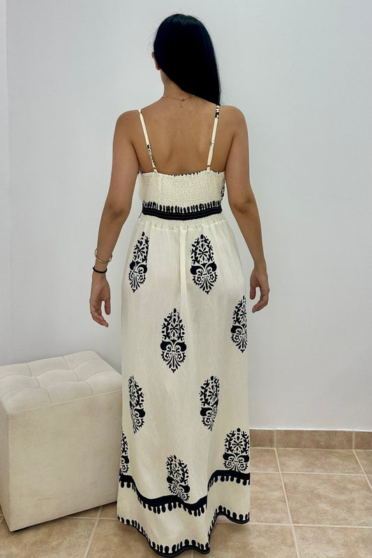 A woman viewed from behind wearing a long white maxi dress with black floral motifs and black trim, standing indoors against a light wall with a small bench and tiled floor visible.