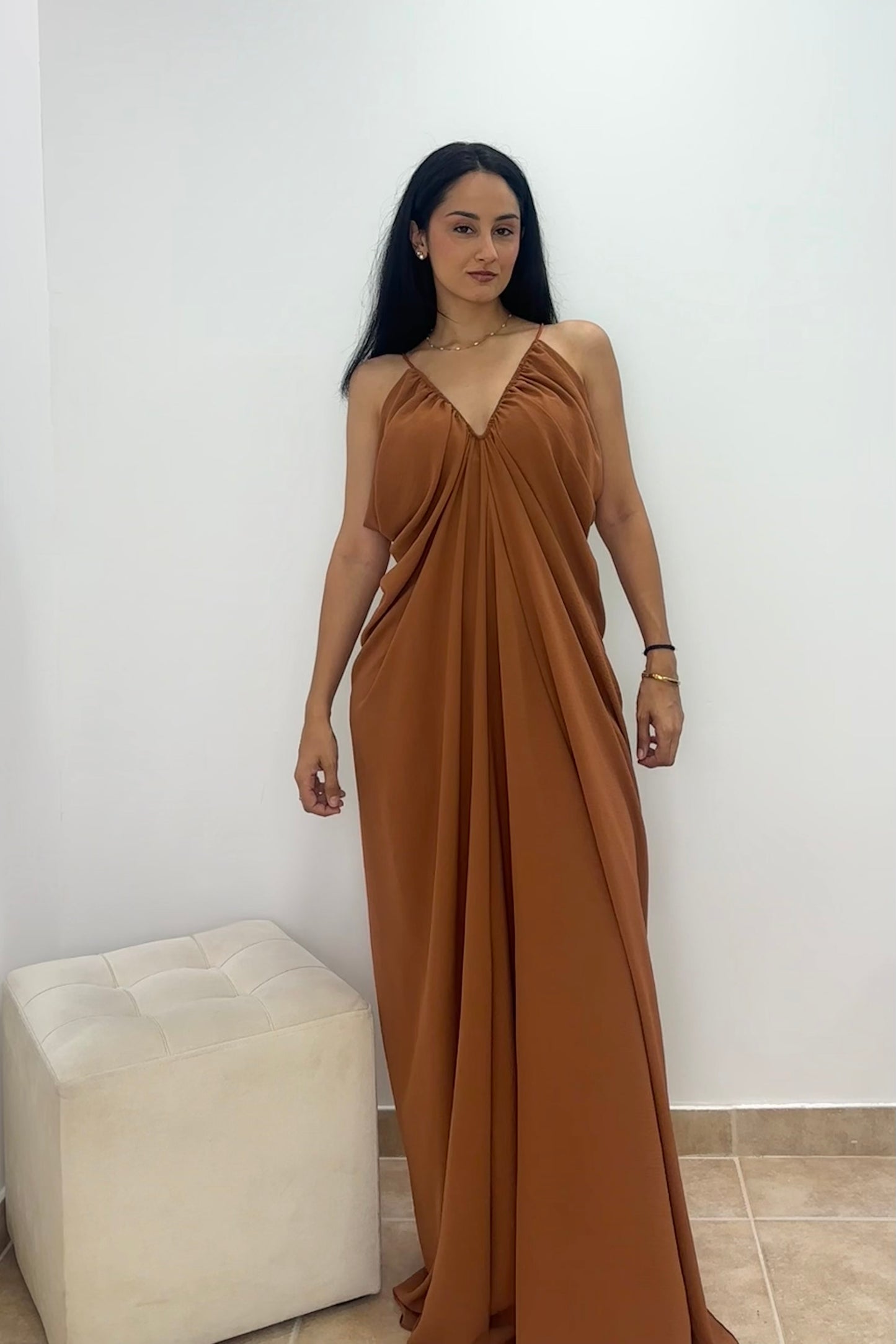 A woman modeling a floor-length draped maxi dress in a warm camel brown color, standing in front of a plain white wall next to a white tufted ottoman.