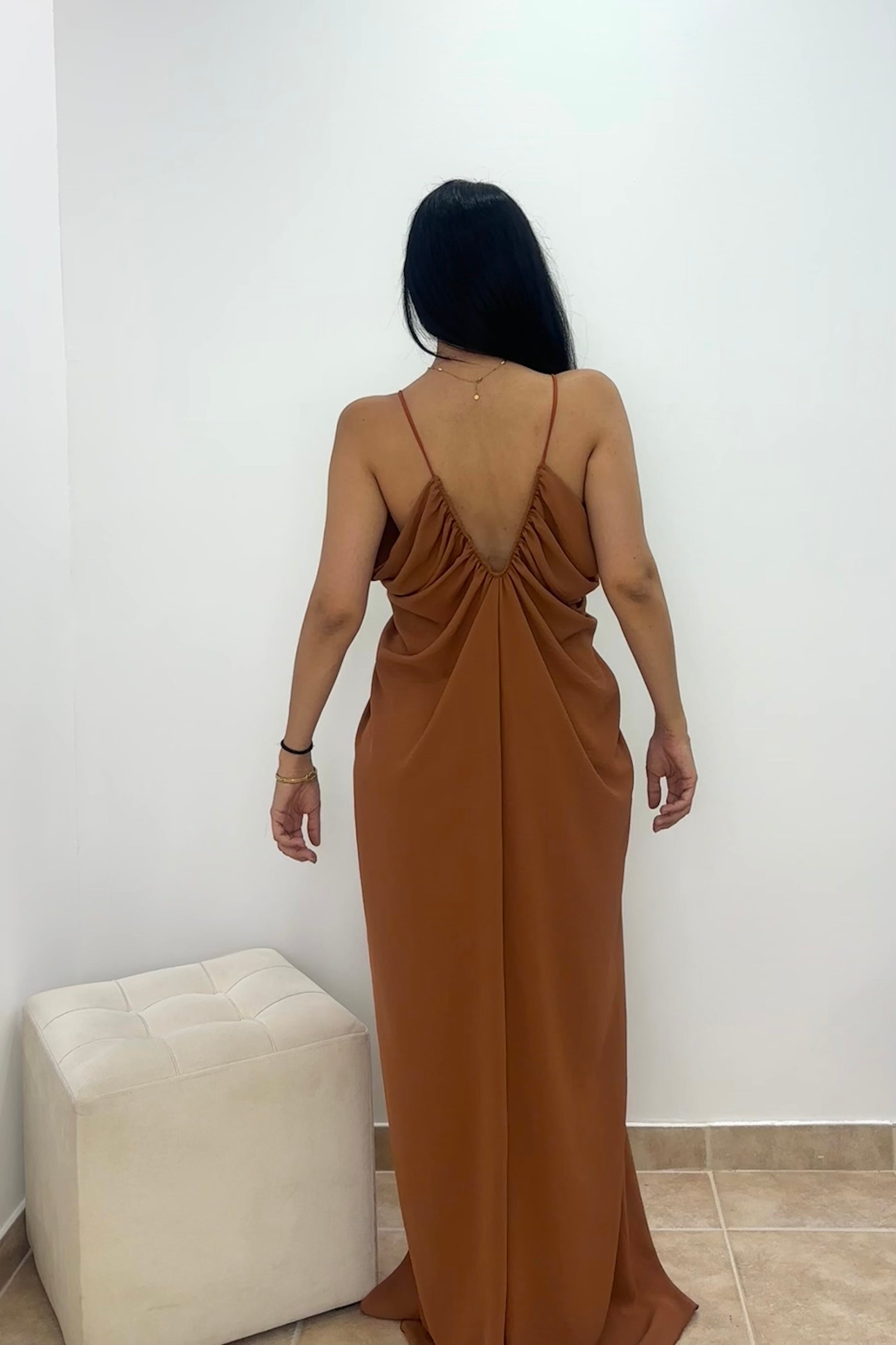 A woman viewed from behind wearing a floor-length draped maxi dress in a warm camel brown color, standing in a minimalist room with a white wall and a cream-colored ottoman.