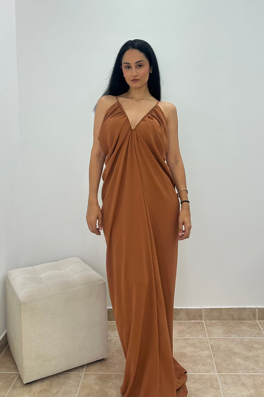 A woman wearing a floor-length draped maxi dress in a warm rust brown color, standing in an indoor setting with a white wall and a light-colored tufted ottoman.