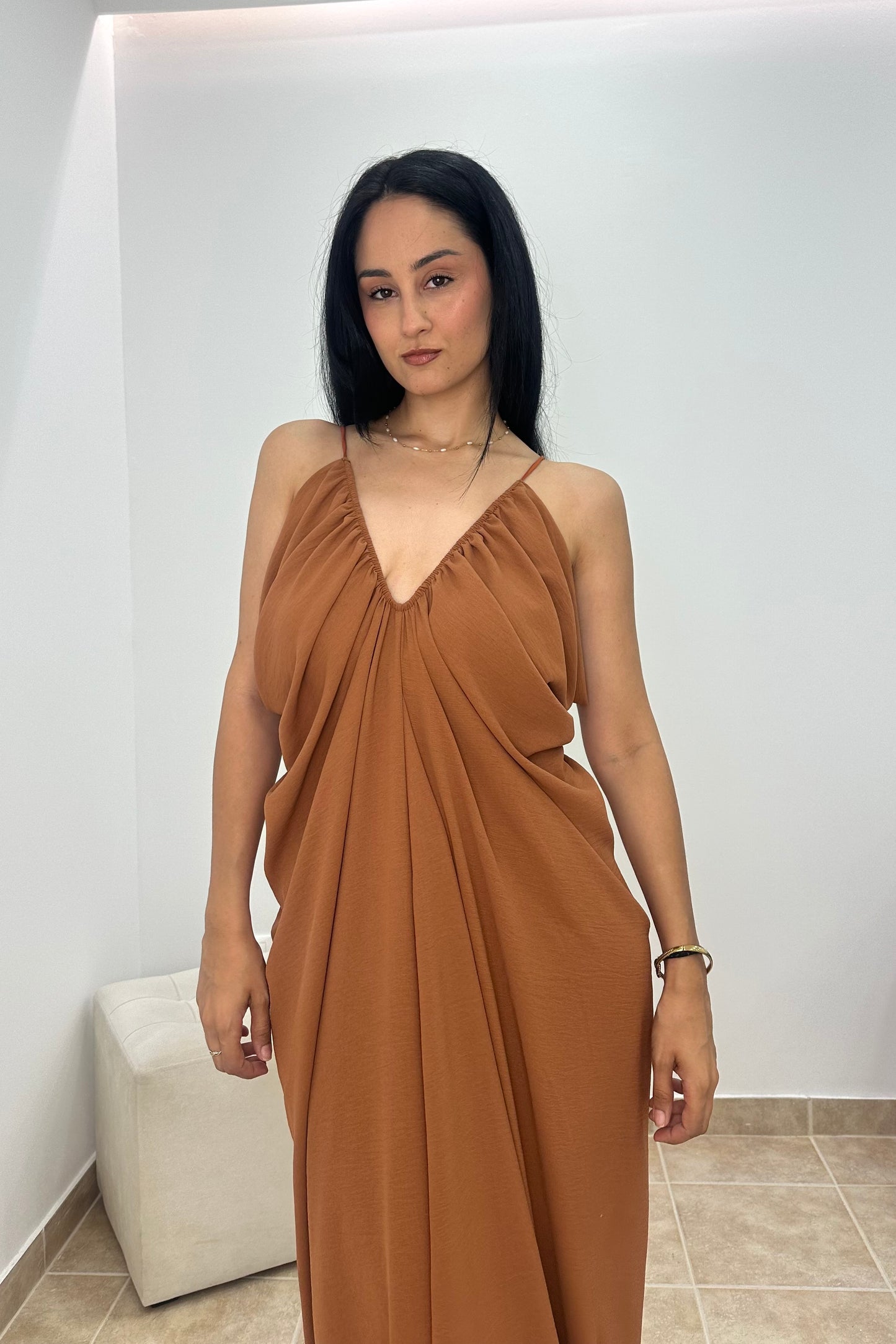 A woman wearing a draped, sleeveless brown maxi dress with a V-neckline, standing in a minimalist room with white walls and tiled flooring.