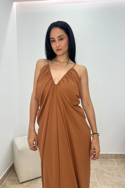 A woman wearing a draped, sleeveless brown maxi dress with a V-neckline, standing in a minimalist room with white walls and tiled flooring.