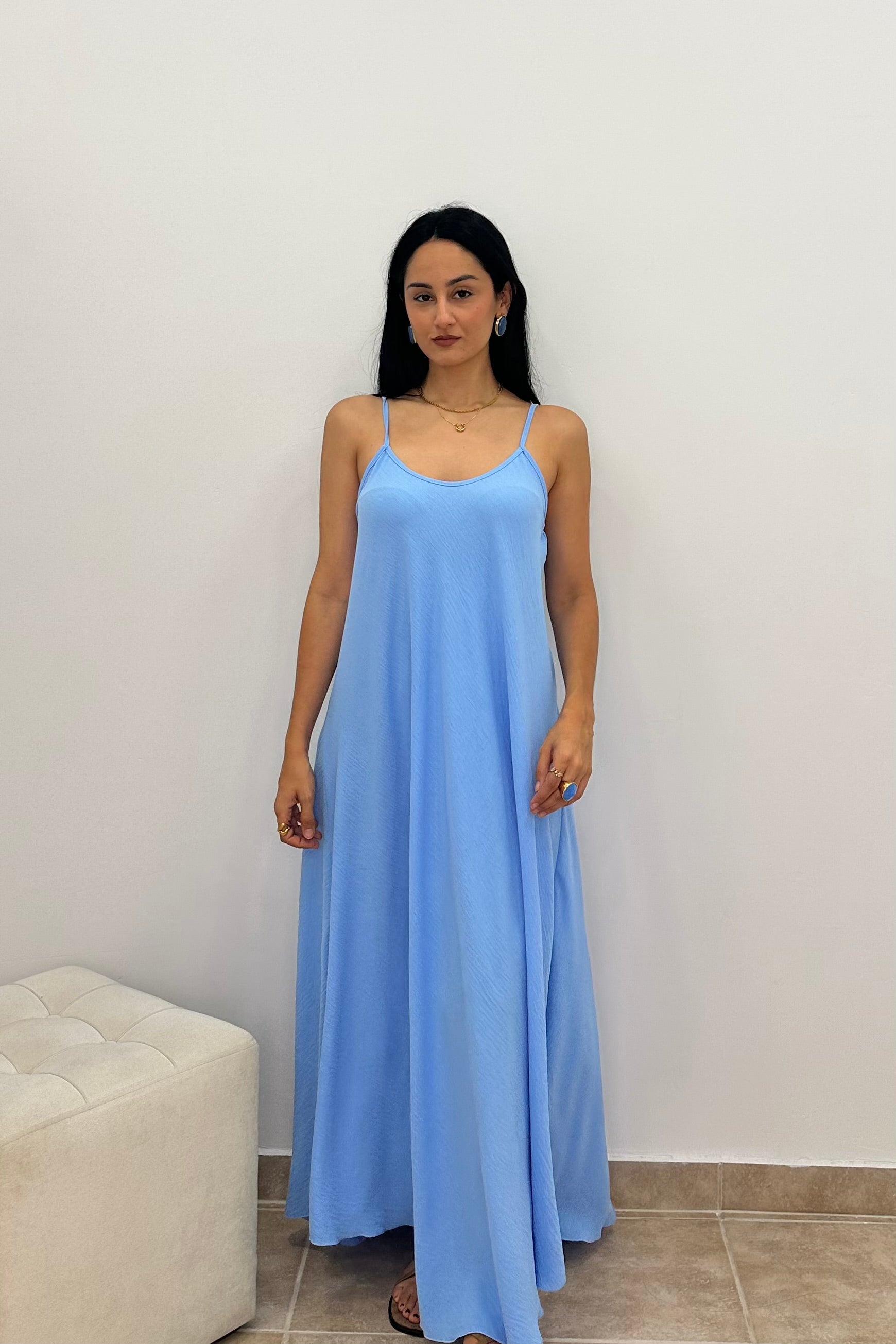 A woman wearing a light blue maxi slip dress with thin spaghetti straps, standing against a plain white wall.
