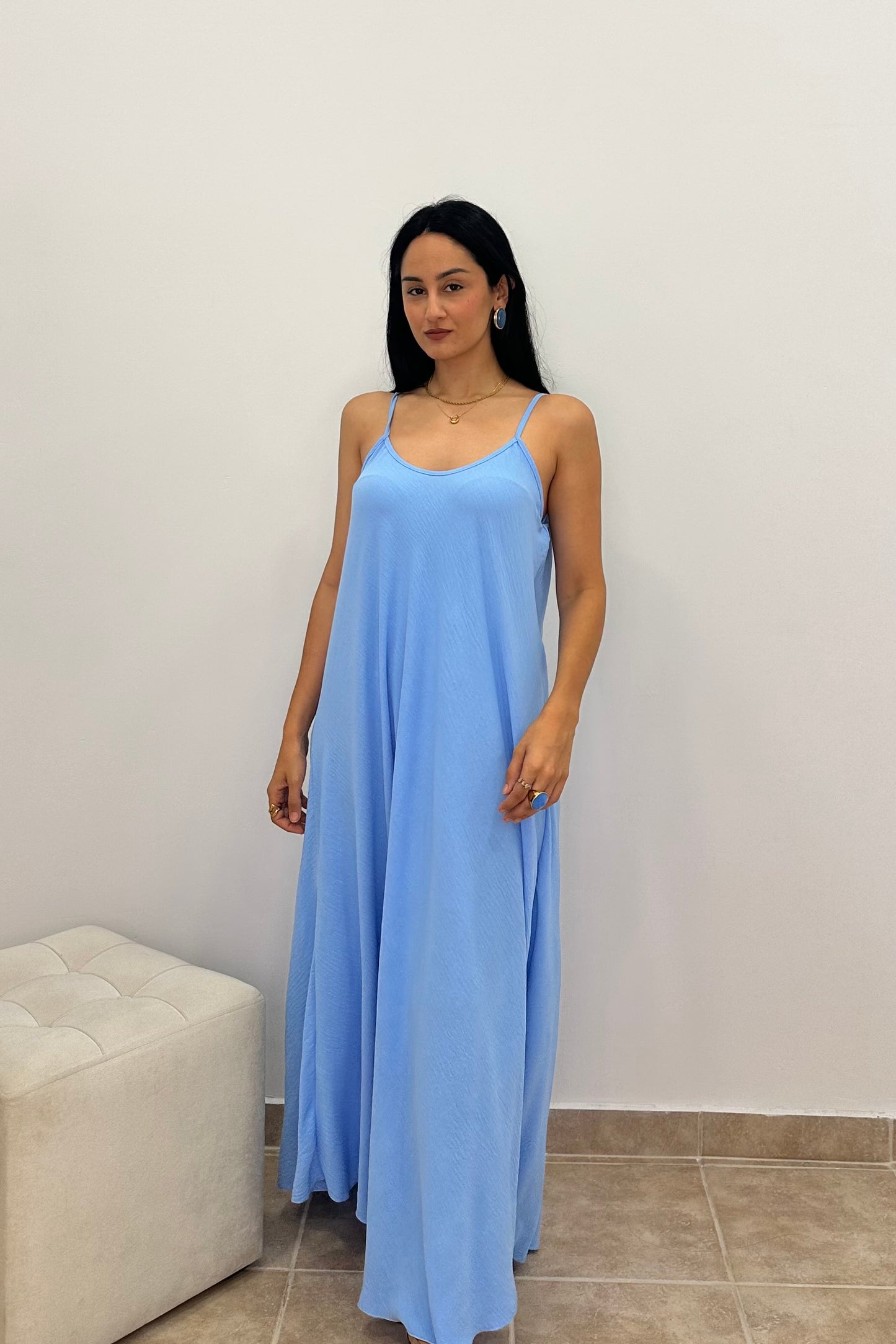 A woman modeling a light blue maxi slip dress with thin spaghetti straps, standing in front of a plain white wall.