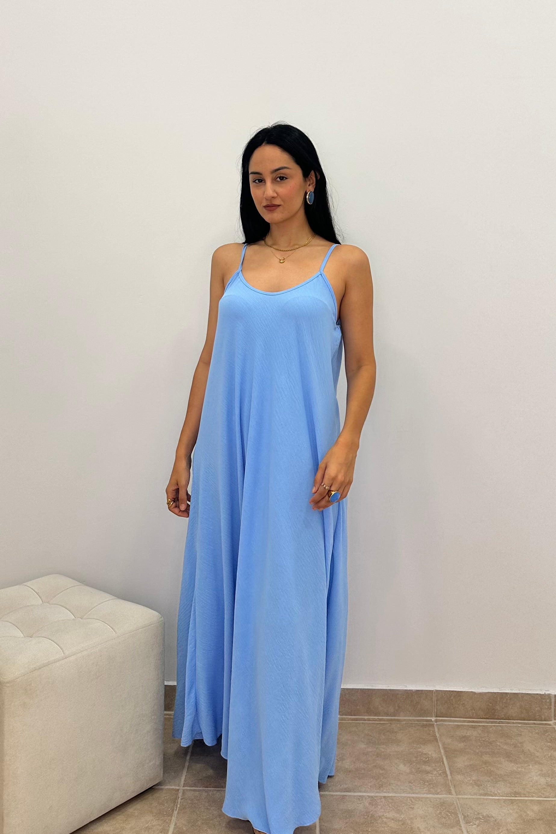 A woman modeling a light blue maxi slip dress with thin spaghetti straps, standing in front of a plain white wall.