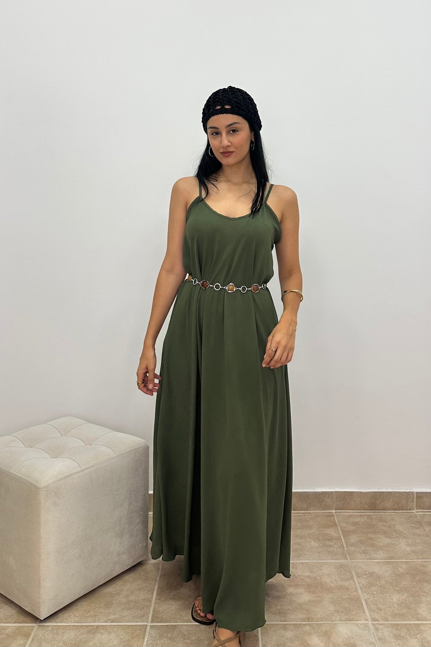 A woman modeling a sleeveless olive green maxi dress with a thin decorative waist belt, standing in a minimalist room with a white wall and a light-colored tufted ottoman.