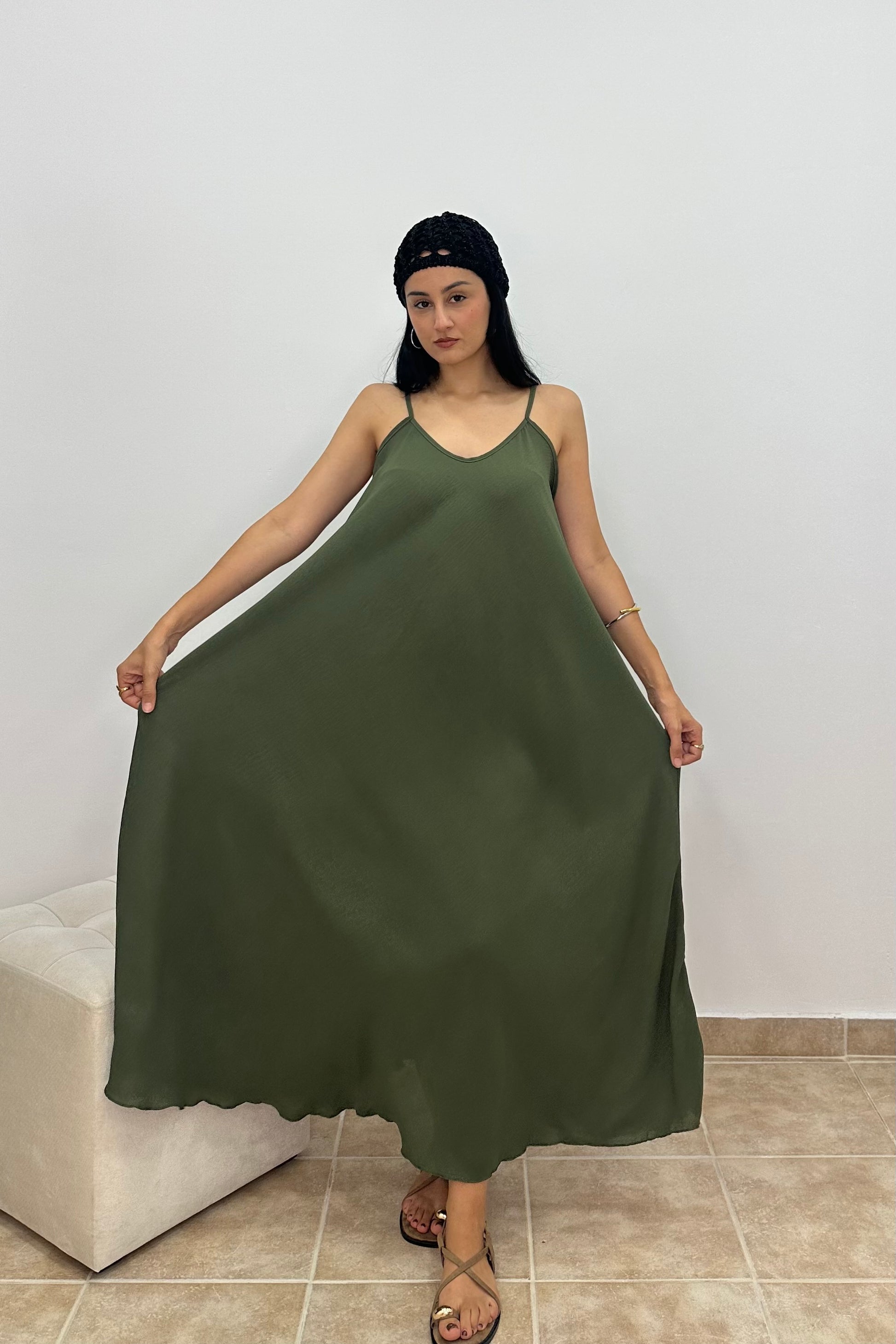 A woman wearing an olive green sleeveless maxi dress with thin straps, holding the skirt out to show its flow. She is standing in a room with a light gray wall and beige tiled floor.