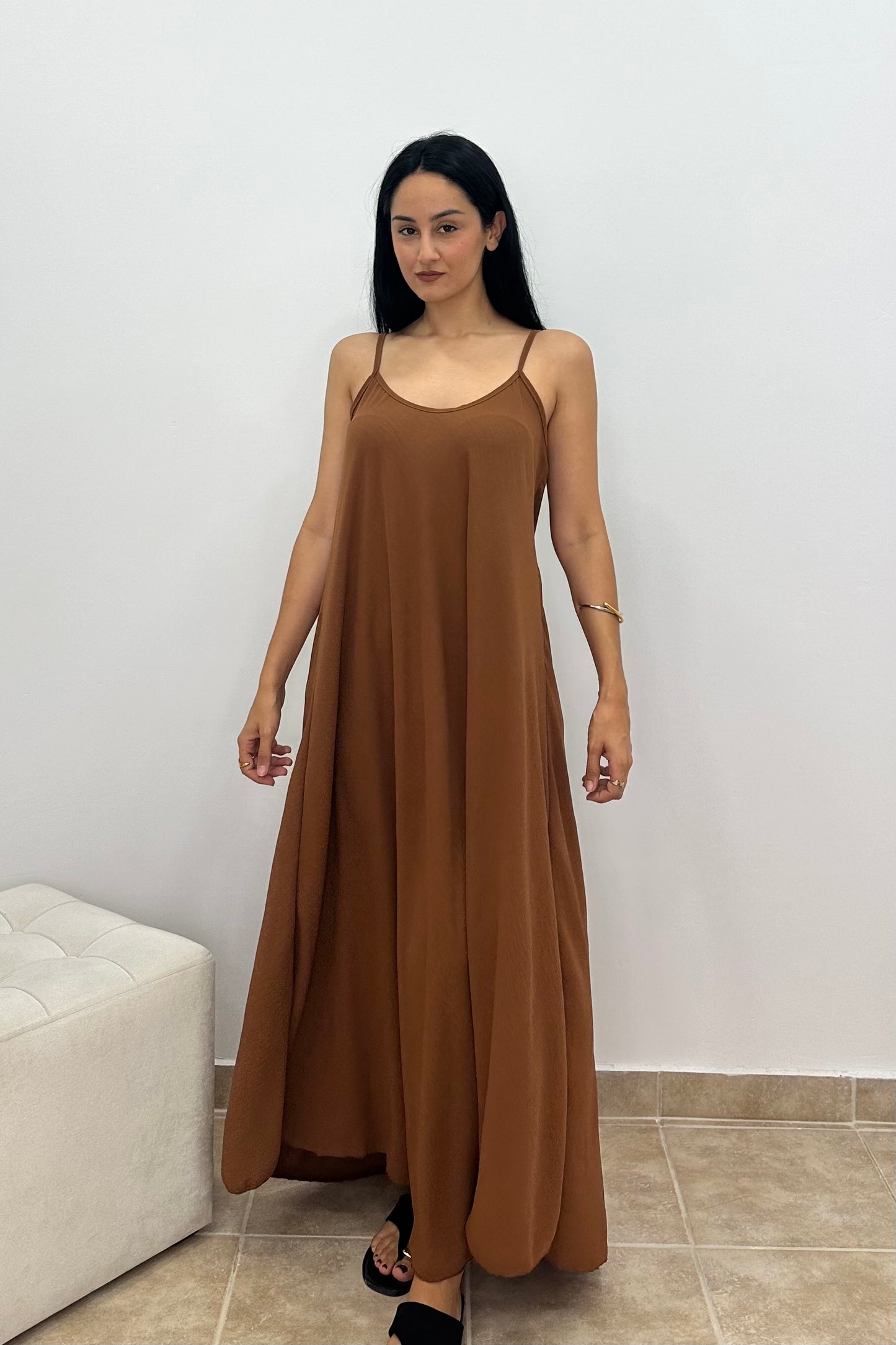 A woman modeling a brown maxi slip dress with thin spaghetti straps, standing in a minimalist room with a white wall and tiled floor.