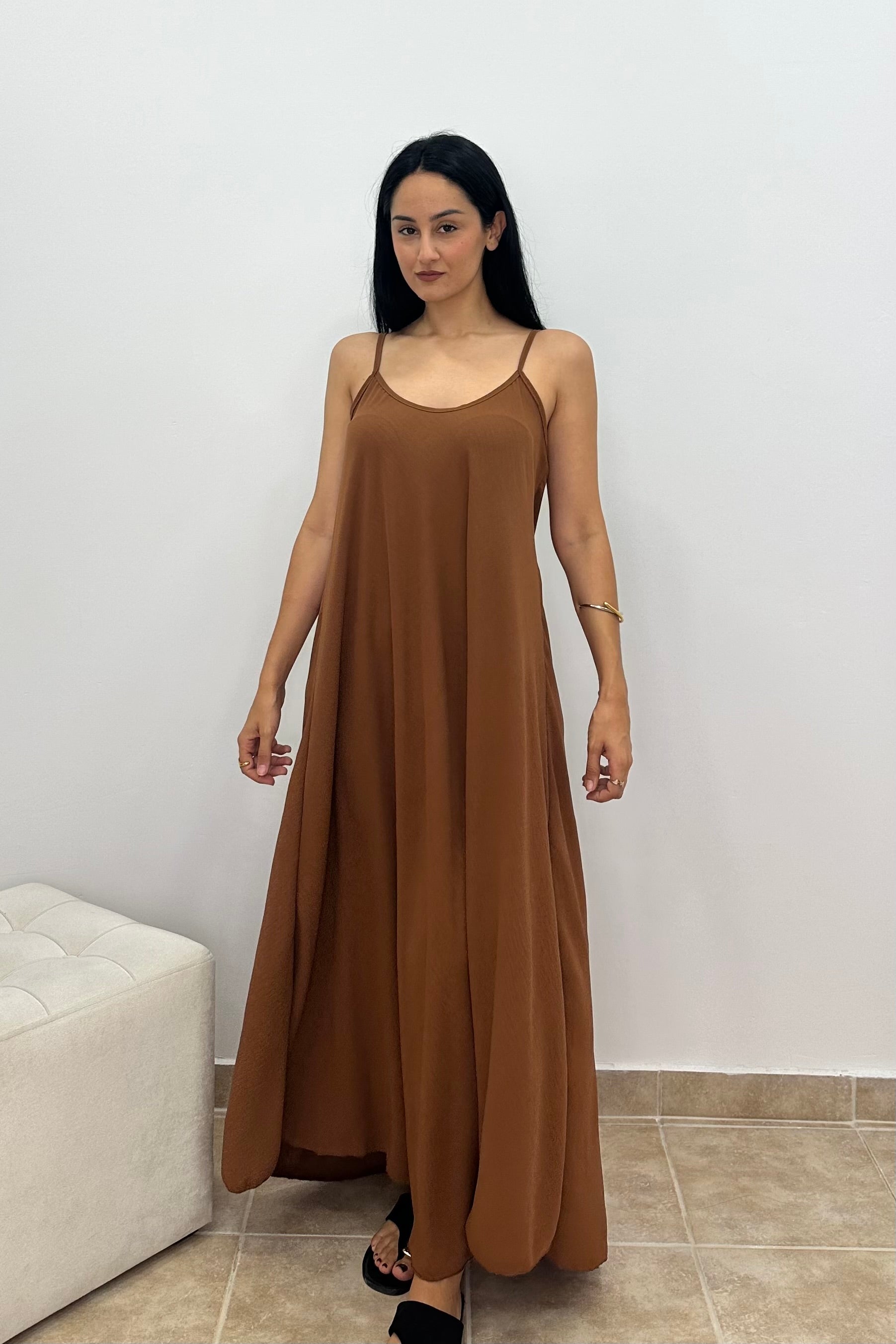 A woman modeling a brown maxi slip dress with thin spaghetti straps, standing in a minimalist room with a white wall and tiled floor.