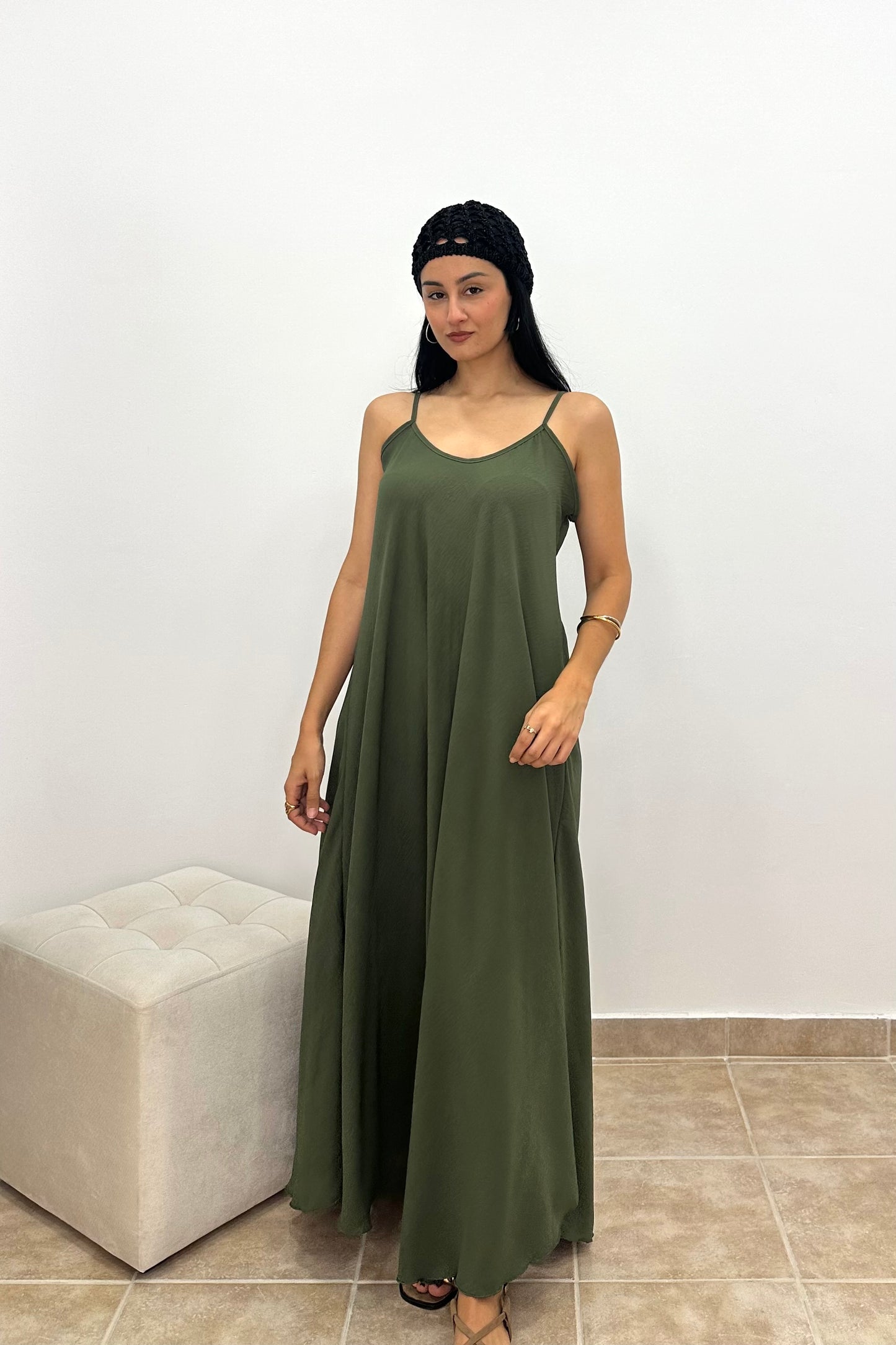 A woman modeling a sleeveless olive green maxi dress with thin spaghetti straps, standing in front of a white wall next to a light beige tufted ottoman.