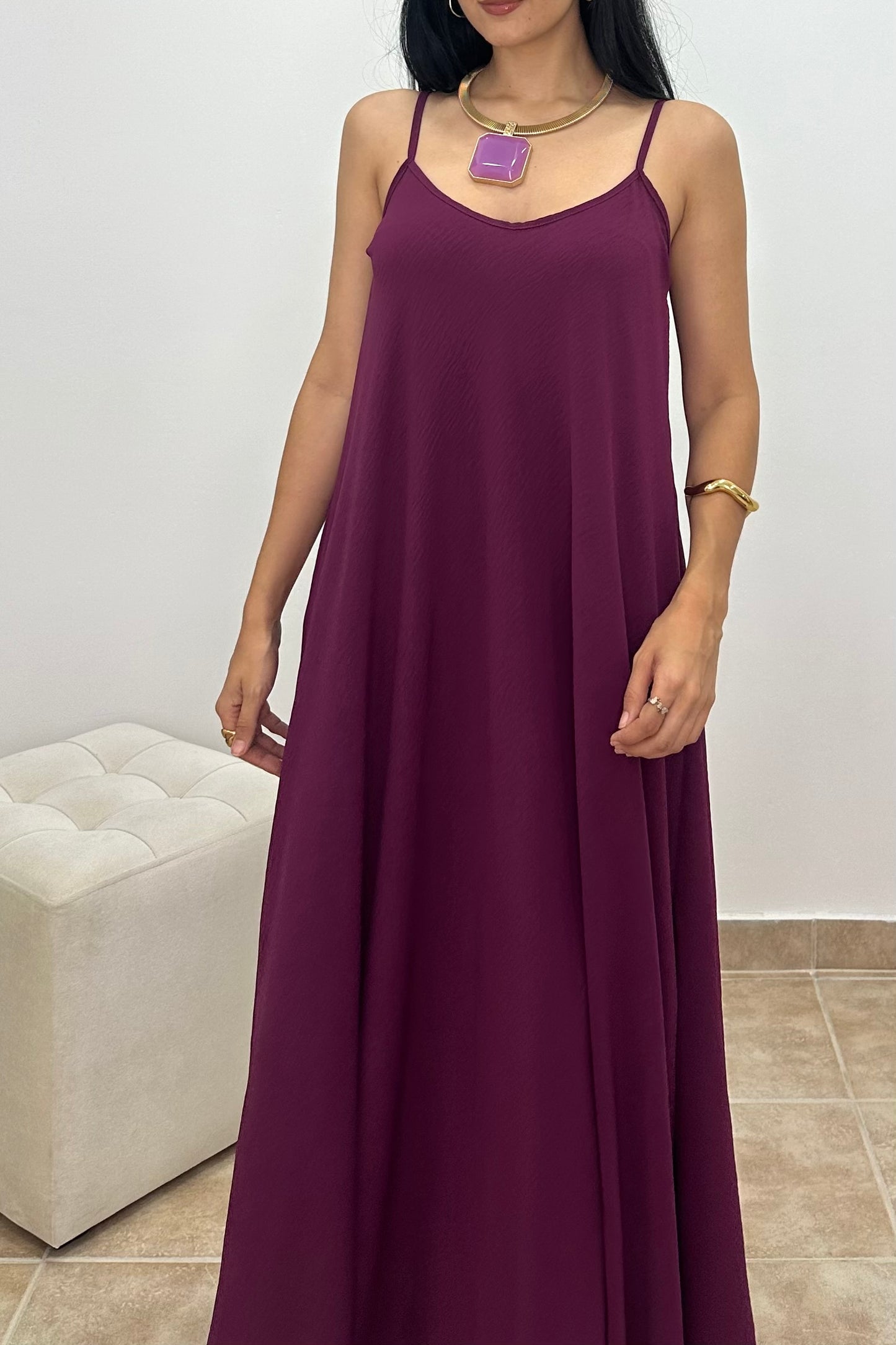 A woman wearing a long, sleeveless, dark purple maxi dress with thin spaghetti straps, standing in an indoor setting with a white tufted ottoman and tiled floor.