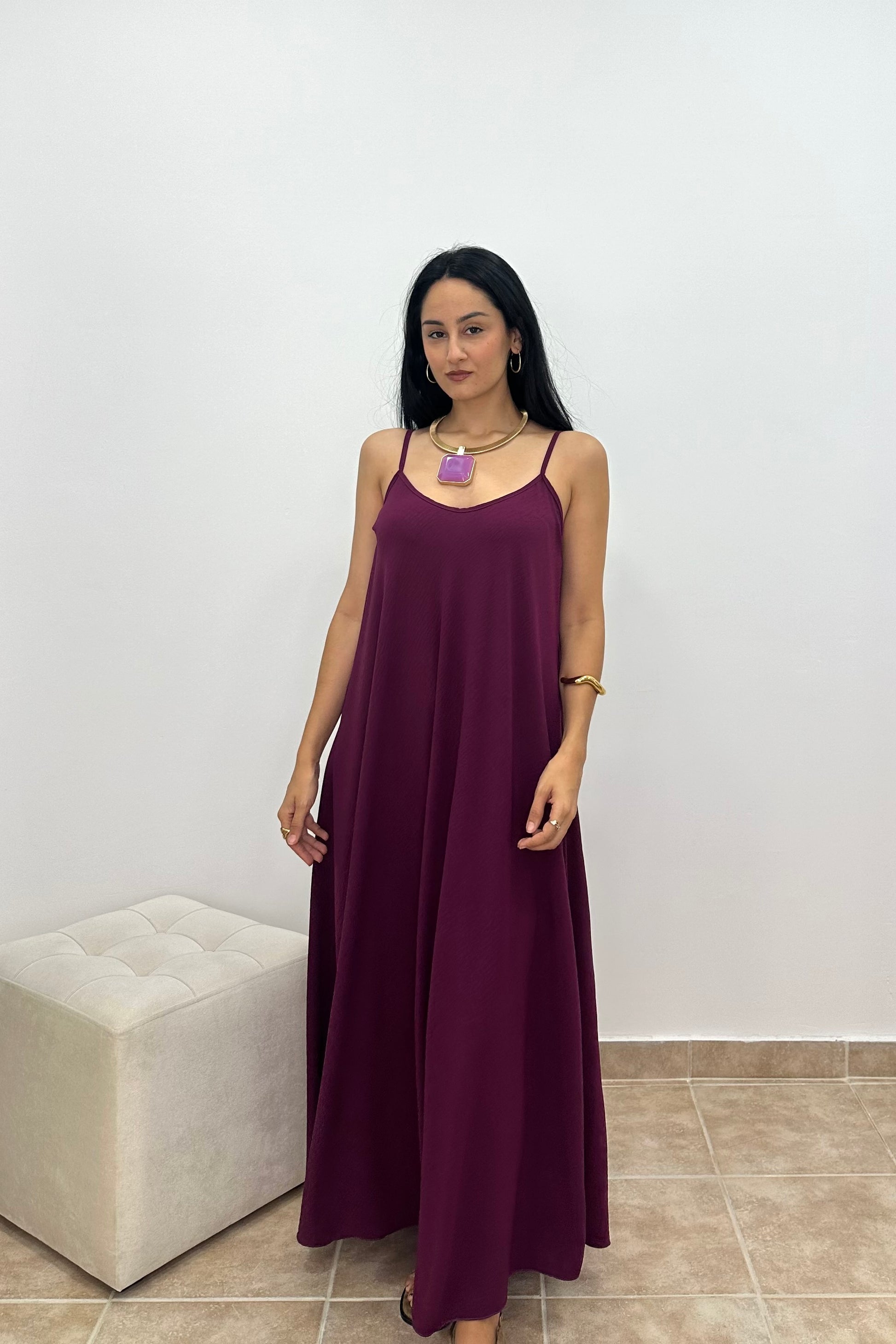 A woman wearing a long, sleeveless, dark purple maxi dress with thin straps, standing in front of a white wall next to a light-colored tufted ottoman.