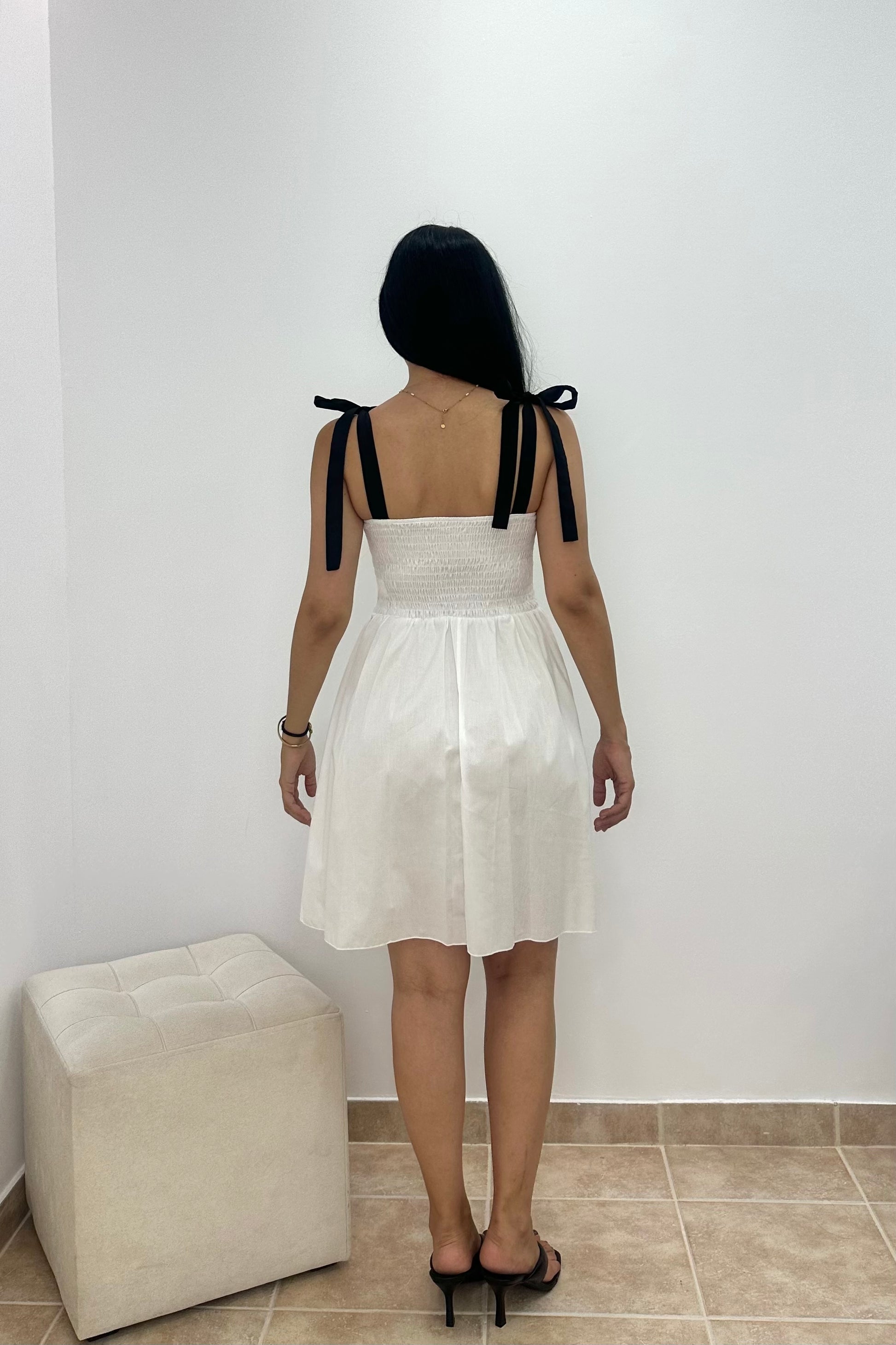 A woman viewed from behind wearing a white sleeveless mini dress with black ribbon tie straps at the shoulders, standing in a room with a white wall and a light-colored ottoman.