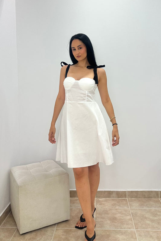 A woman modeling a white strapless mini dress with black shoulder ties, standing in a minimalist room with a light-colored floor and a tufted ottoman.