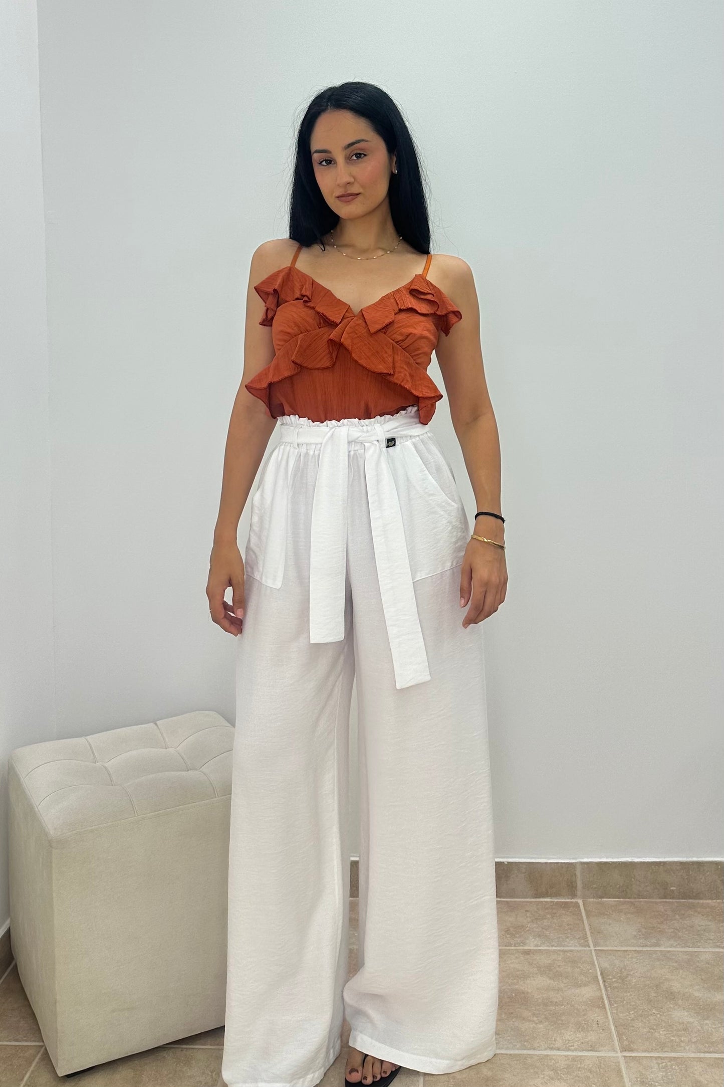 A woman modeling a two-piece outfit consisting of a rust-orange ruffled strapless top and high-waisted white wide-leg pants with a matching tie belt, standing in a minimalist room with a white wall and a light-colored ottoman.