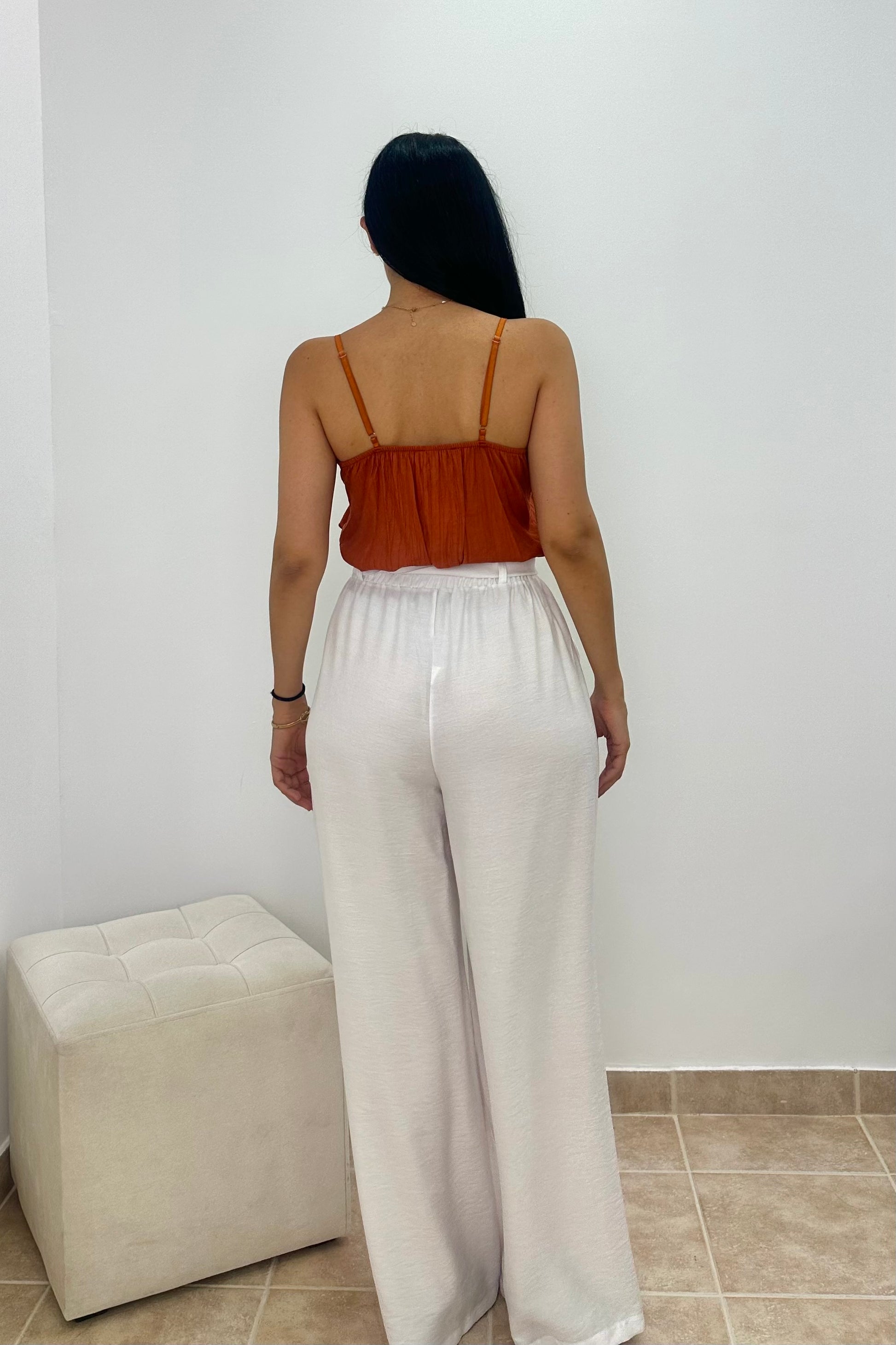 A woman shown from the back wearing a rust-orange spaghetti strap top and white wide-leg pants, standing in a room with a white wall and a beige ottoman.