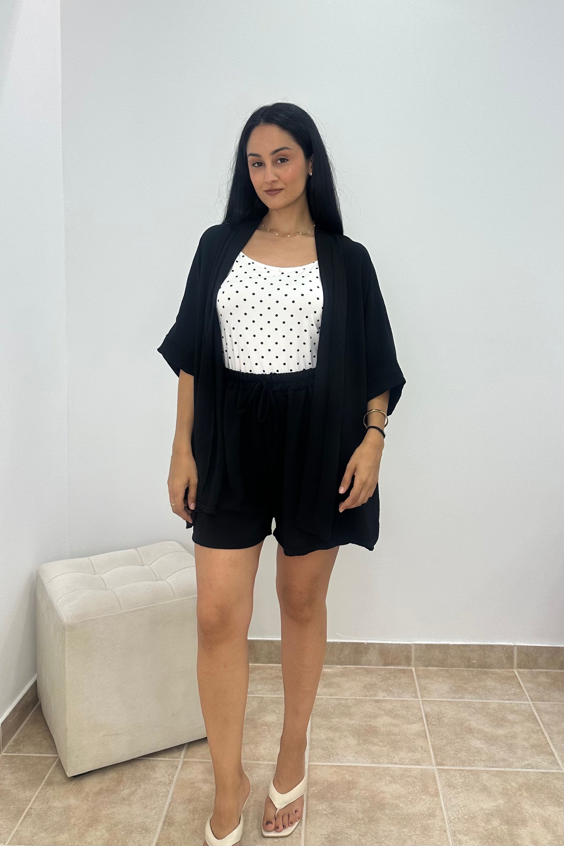 A woman modeling a black short-sleeve open-front cardigan over a white sleeveless top with black polka dots and black shorts, standing in a room with a light gray wall and beige tiled floor.