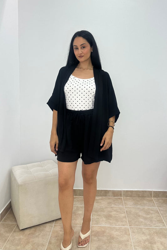 A woman modeling a black short-sleeve open-front cardigan over a white sleeveless top with black polka dots and black shorts, standing in a room with a light gray wall and beige tiled floor.