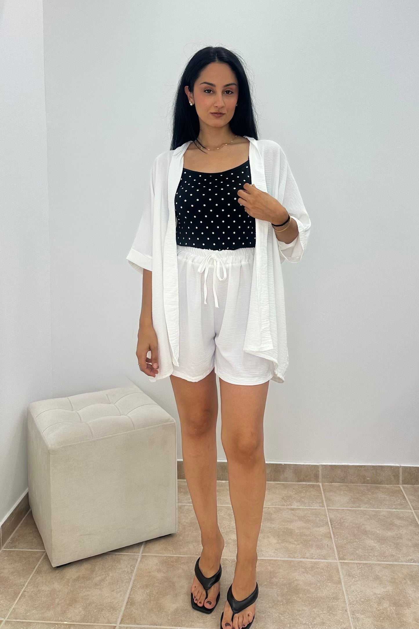 A woman modeling a white short-sleeve open-front cardigan over a black and white polka dot top and white shorts, standing indoors against a white wall with a light-colored ottoman and tiled floor.